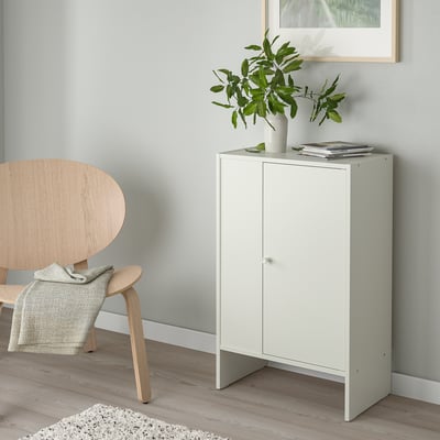 Bílá úložná skříňka s časopisy a rostlinami. Dřevěné křeslo s dekou, minimalistický dekor.