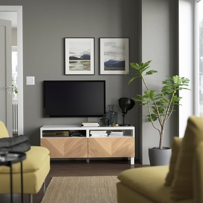 Moderní obývací pokoj s bílým TV stolkem BESTÅ, zásuvkami se vzorem šípů, TV s plochou obrazovkou, žlutým křeslem a vysokou zelenou rostlinou.