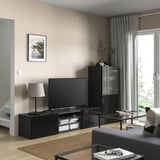 Moderní obývací pokoj s černou TV stolkem BESTÅ, TV s plochou obrazovkou, šedými závěsy a šedým kobercem na světlé dřevěné podlaze.