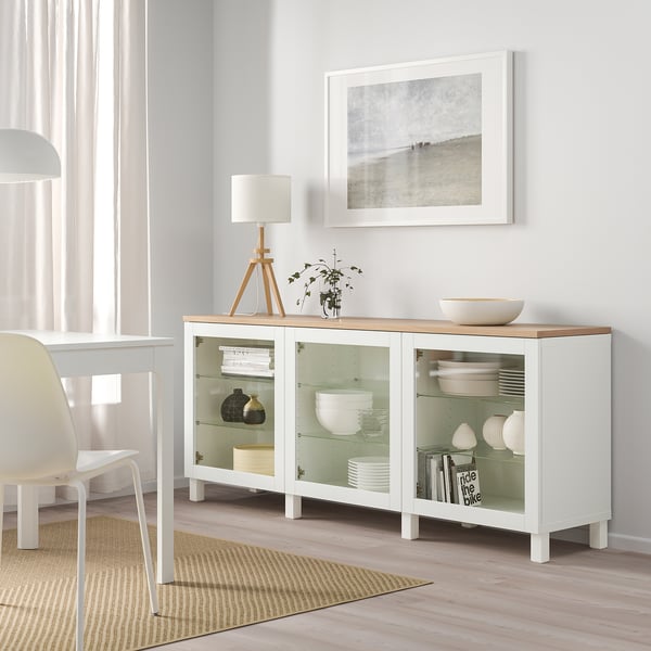 Bílý příborník ikea BESTÅ se skleněnými dveřmi, vystavující nádobí a vázy, spolu s lampou, rostlinou a obrázkem pláže ve světlé jídelně.