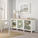 Bílý příborník ikea BESTÅ se skleněnými dveřmi, vystavující nádobí a vázy, spolu s lampou, rostlinou a obrázkem pláže ve světlé jídelně.