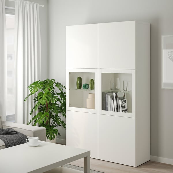 Velká bílá skříňka BESTÅ se skleněnými dvířky, na kterou lze uložit dekorativní předměty. Moderní a minimalistický design, umístěný vedle velké rostliny.