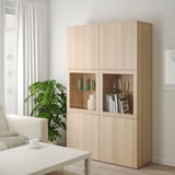 Ikea BESTÅ úložná skříňka ze světlého dubu se skleněnými dvířky a smíšenými policemi vedle konferenčního stolku a pohovky.
