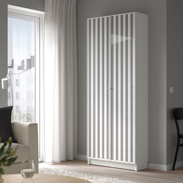 Vysoká, bílá knihovna BILLY se skleněnými dvířky stojí vysoko vedle okna. Moderní, minimalistický design.