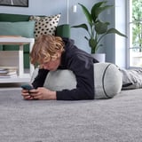 Dítě ležící na šedém polštáři BLÅSKATA, používající chytrý telefon na podlaze s kobercem, se zelenou pohovkou a rostlinou poblíž.