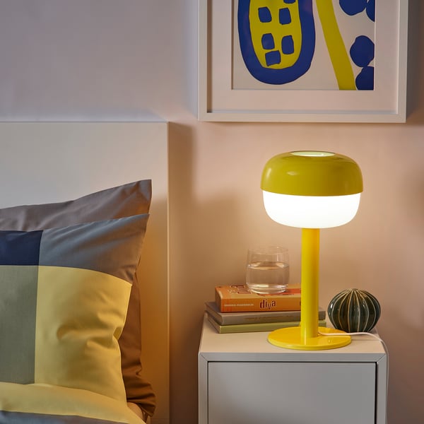 Jasně žlutá lampa BLÅSVERK na nočním stolku, malé sklo a knihy. Moderní retro design.