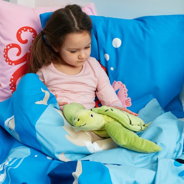 Dívka objímá měkkou zelenou želvu v posteli pod modrou dekou s motivem mořských tvorů.