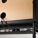 Černý TV stolek s nastavitelnými nožičkami a spodní síťkou na kabely, připevněný ke stěně z korkové nástěnky.