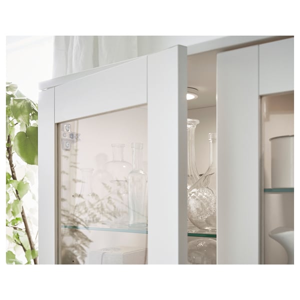 Bílá vitrína se skleněnými dveřmi, kde jsou uloženy vázy z čirého skla, opřená o bílou stěnu. Lehký, moderní design.