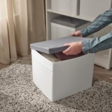 Osoba otevírající šedé látkové víko na bílém úložném boxu na podlaze s kobercem.