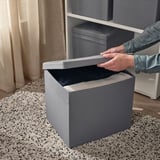 Osoba otevírá šedé víko krabice DRÖNA na kobercové podlaze vedle bílého regálu.