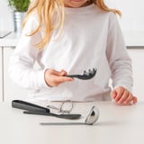 Dítě držící servírku na špagety s dalšími kuchyňskými potřebami na stole, což podporuje předstíranou hru na vaření.