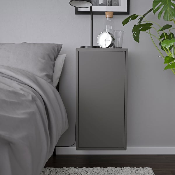 Moderní šedá nástěnná skříňka, součást série IKEA EKET, s hodinami a lampou, namontovaná vedle postele.