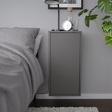 Moderní šedá nástěnná skříňka, součást série IKEA EKET, s hodinami a lampou, namontovaná vedle postele.