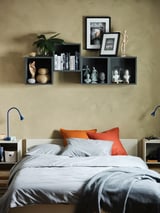 Moderní ložnice s béžovými stěnami, šedou postelí s oranžovými polštáři a černými nástěnnými úložnými kostkami EKET s knihami, rostlinami a dekoracemi.