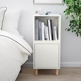 Moderní ložnice s bílým nočním stolkem vedle postele, držícím knihy, budík a rostlinu. Počet znaků: 44