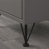 Tmavě šedý úložný díl na dřevěné podlaze s tenkými černými kovovými nohami pro stabilitu a elegantním designem.