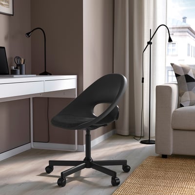 Moderní domácí kancelář s černou židlí ELDBERGET, elegantním designem, polstrovaným sedákem, nastavitelnou výškou, kolečky, téměř bílým stolem a šedými závěsy.