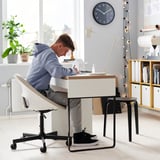 Muž sedící u moderního stolu se židlí ELDBERGET, kreslí. Bílá židle, světlý dřevěný stůl, lampa a knihovna. Hodiny na zdi.