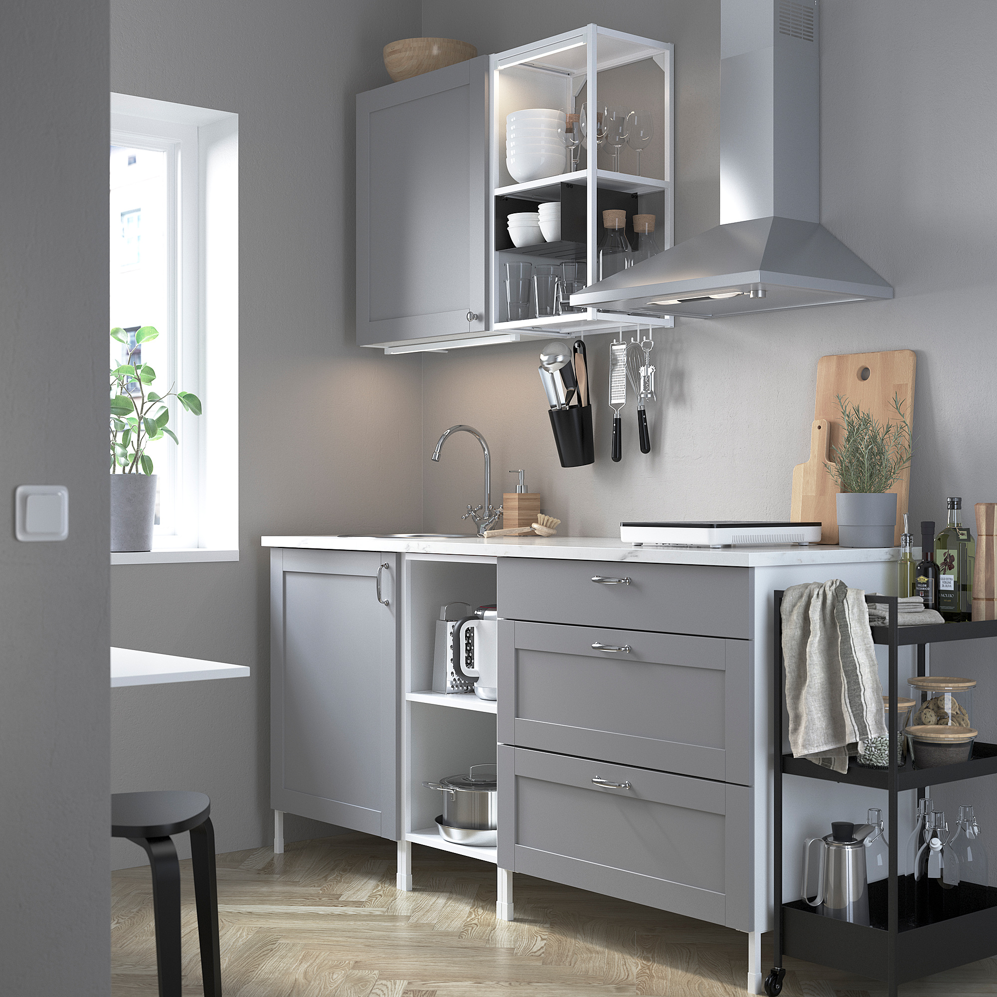 Moderní šedá kuchyňská linka ENHET s elegantním úložným prostorem, dřezem a spotřebiči. Rostliny a desky zvyšují efektivitu prostoru.