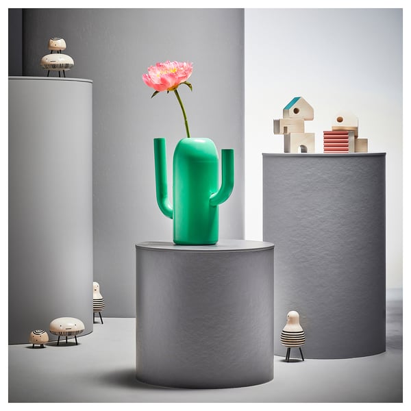 Zelená váza ve tvaru kaktusu s růžovým květem na šedém podstavci. Dřevěné figurky a bloky v okolí. Moderní minimalistický design.