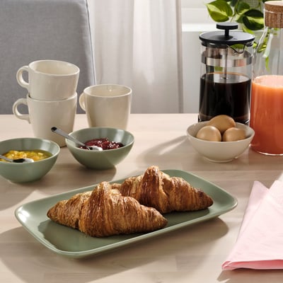 Snídaňový stůl s talířem croissantů, mísami s ovocem a džemem, hrnky a lisem na kávu, kde je vystaveno nádobí FÄRGKLAR.