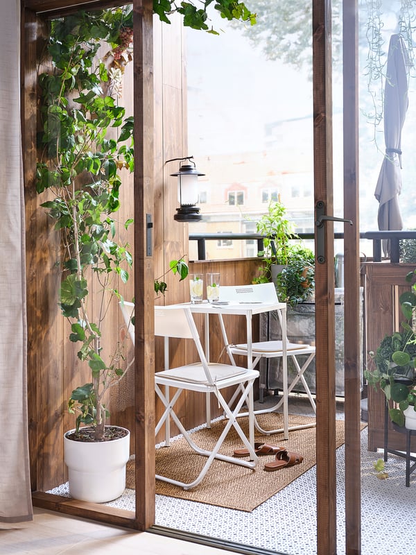 Malý balkon s bílými skládacími židlemi, stolem a rostlinami v květináčích. Dřevěná podlaha, prosklené dveře.