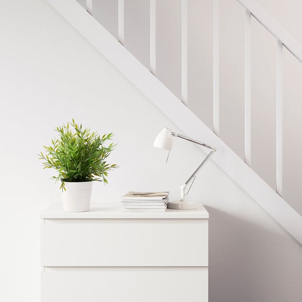 Bílé schodiště s bílou skříňkou pod ním. Je v něm lampa, hromada knih a zelená rostlina v květináči.