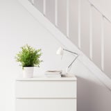 Bílé schodiště s bílou skříňkou pod ním. Je v něm lampa, hromada knih a zelená rostlina v květináči.