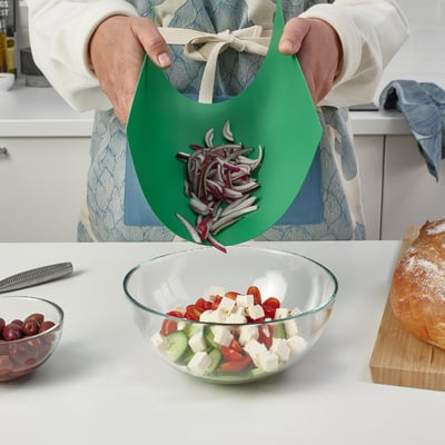 Osoba držící pružné prkénko s nakrájenou cibulí a zasouvá je do salátové mísy obsahující rajčata, okurky a sýr.