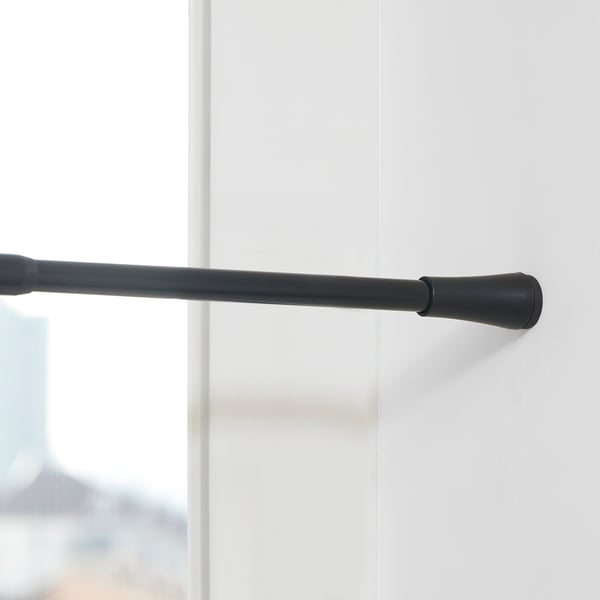 Černá nastavitelná tyč na závěsy namontovaná na bílé stěně, sahající od 70 do 120 cm, s gumovými krytkami na koncích, které zabraňují poškrábání.