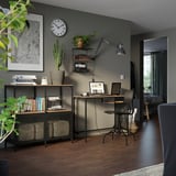 Moderní domácí pracovna s policí, psacím stolem a židlí FJÄLLBO. Podlaha z tmavého dřeva, šedé stěny, rostliny a hodiny. Industriální a rustikální design.
