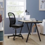 Nastavení domácí kanceláře s černou ergonomickou síťovanou židlí, dřevěným stolem, notebookem, lampou a stoličkou u modré stěny.