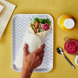 Osoba držící wrap se zeleninou na vzorovaném podnosu vedle sklenice s okurkami a žlutého šálku.