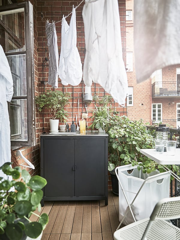 Balkon s dřevěnou podlahou, cihlovou stěnou, věšeným prádlem. Tmavá skříňka, rostliny v květináčích a židle.