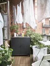 Balkon s dřevěnou podlahou, cihlovou stěnou, věšeným prádlem. Tmavá skříňka, rostliny v květináčích a židle.