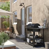 Moderní venkovní gril s černým kovovým grilem na dřevěné uhlí GRILLSKÄR, obklopený příslušenstvím na dlážděné terase u vchodu do domu.