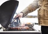 Osoba griluje jídlo na grilu na dřevěné uhlí GRILLSKÄR s černým smaltem a nerezovými akcenty, venkovní prostředí.