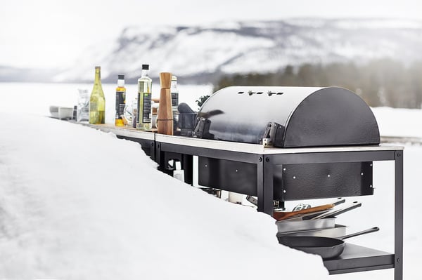Zimní grilovací sestava s grilem GRILLSKÄR, obklopená oleji a kořením, zasněžená horská kulisa.