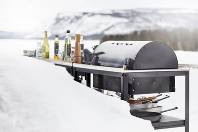Zimní grilovací sestava s grilem GRILLSKÄR, obklopená oleji a kořením, zasněžená horská kulisa.