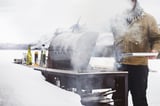 Muž griluje v zasněženém dni pomocí stříbrného grilu GRILLSKÄR se stoupající párou, držící bílý talíř, obklopený lahvemi.