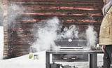 Osoba griluje venku pomocí černého grilu GRILLSKÄR a vydává kouř. Sníh pokrývá zem a v pozadí je dřevěná chatka.