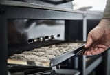 Osoba manipulující s černým smaltovaným grilem GRILLSKÄR s nerezovými díly, vysouvacím popelníkem.