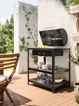 Moderní venkovní gril s grilem GRILLSKÄR, černý, nerezová ocel, obdélníkový, na dlážděné terase, obklopený zelení.