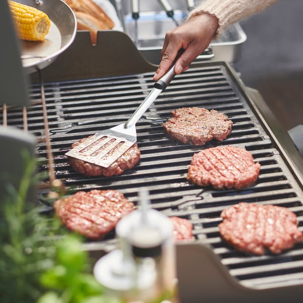 Osoba grilující hamburgery. Používají nástroje GRILLTIDER, stříbrné a černé, na grilu.