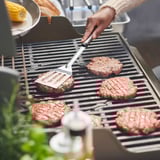 Osoba grilující hamburgery. Používají nástroje GRILLTIDER, stříbrné a černé, na grilu.