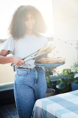 Žena držící modrý talíř s chlebem, používající stěrku GRILLTIDER, na terase s rostlinami, na sobě bílou košili a džíny.