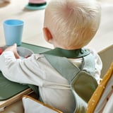Dítě v zelenobílém bryndáku GULDVÄVARE, navrženém s kapsou na chytání jídla, sedící na vysoké židličce, jící.