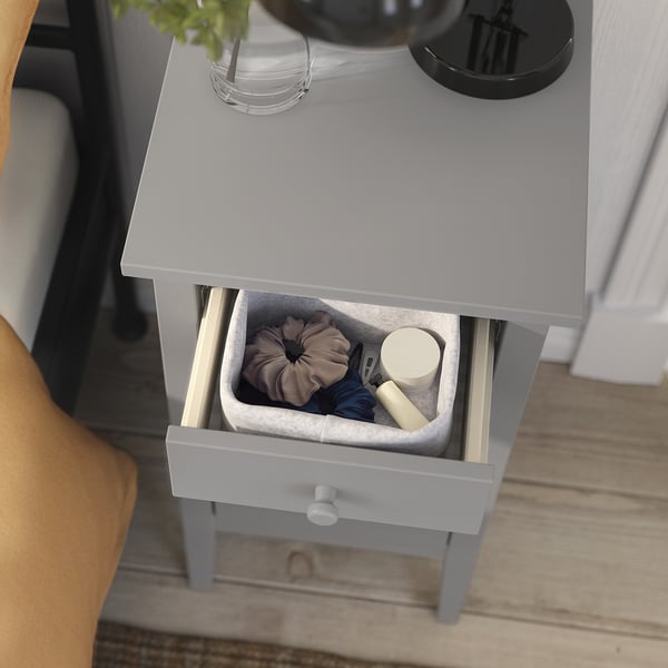 Šedý noční stolek gullberg s košíkem na vlasové doplňky, vedle bílého radiátoru na podlaze ze světlého dřeva.