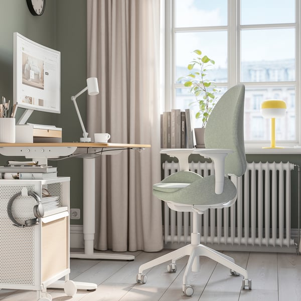 Moderní zelená kancelářská židle na minimalistickém stole s lampou, počítačem a rostlinami u okna.